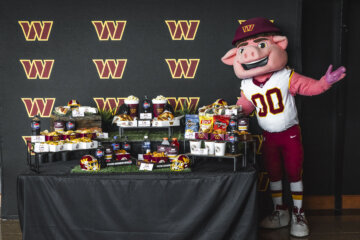 Washington Commanders' mascot, a giant anthropomorphic pig, poses to viewers' right of concessions arranged on a table at a press conference