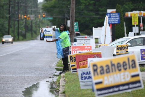 man greeting voters by street of polling place with campaign signs in foreground
