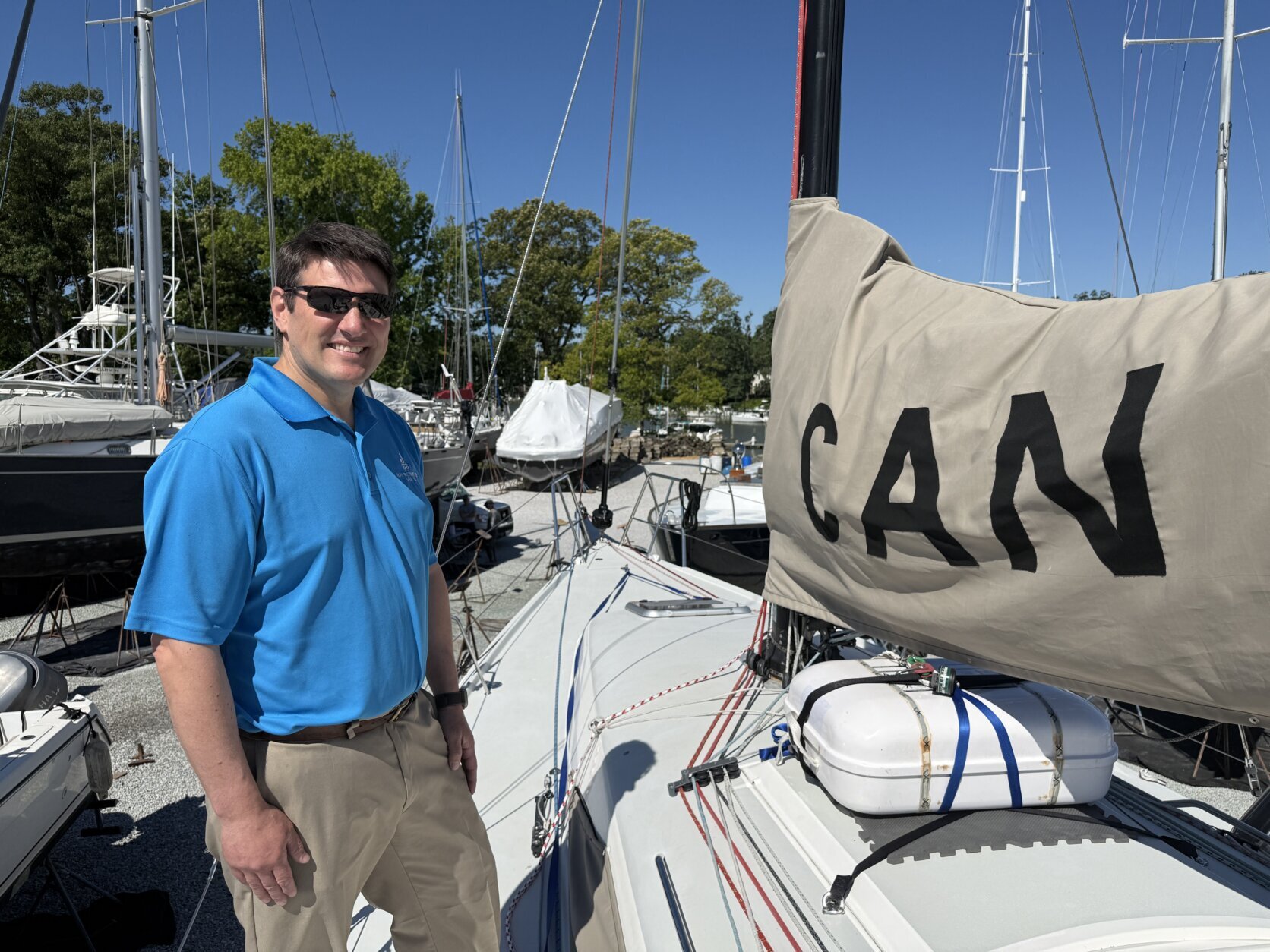 a man in blue polo smiles at the camera from viewer's left of a sailboat.