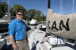 a man in blue polo smiles at the camera from viewer's left of a sailboat.