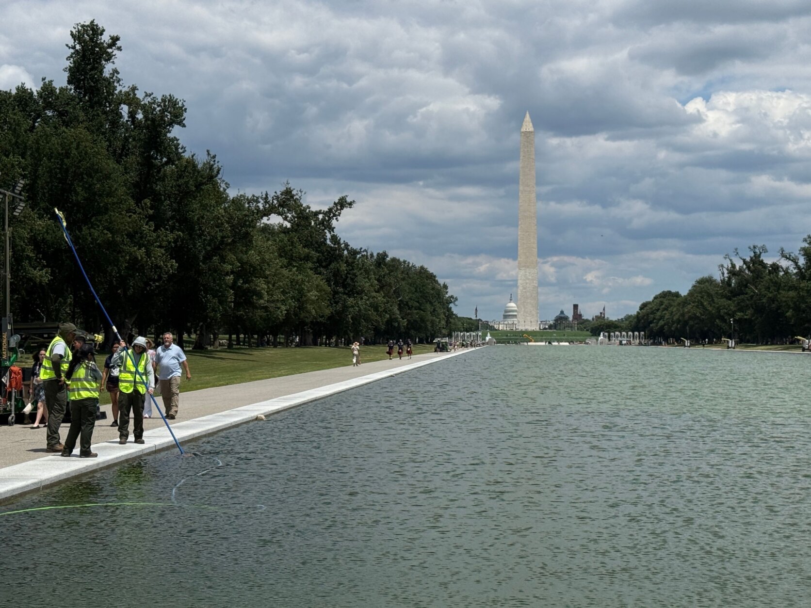 Workers in high-viz vests next to the reflecting pool. The Washington Monument towers in the distance.