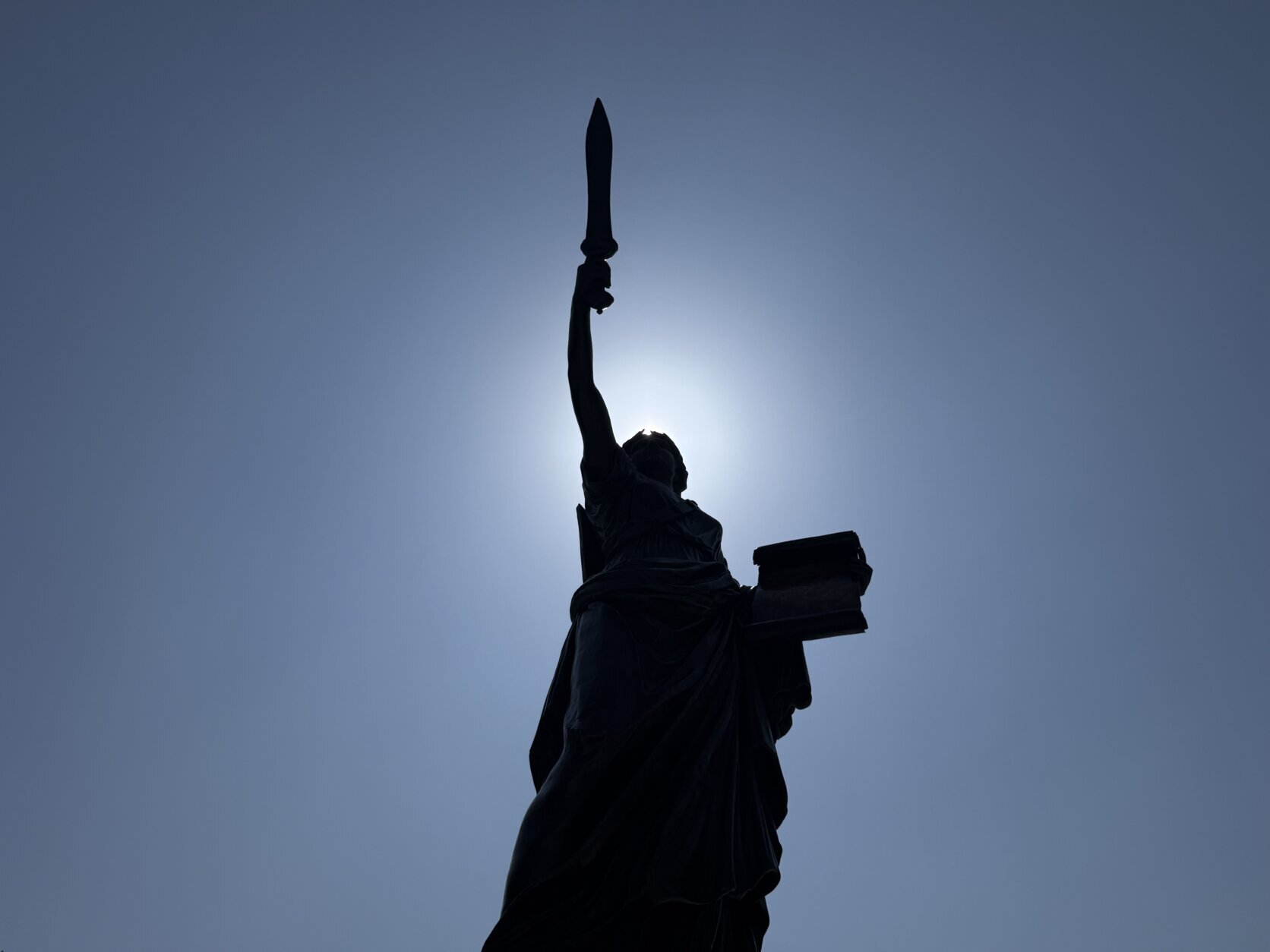 statue holding torch pictured from below with blue sky and sun silhouetting statue