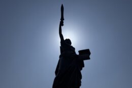 statue holding torch pictured from below with blue sky and sun silhouetting statue