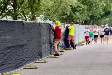 Workers cover a chain link fence on the National Mall with black material as tourists look on.