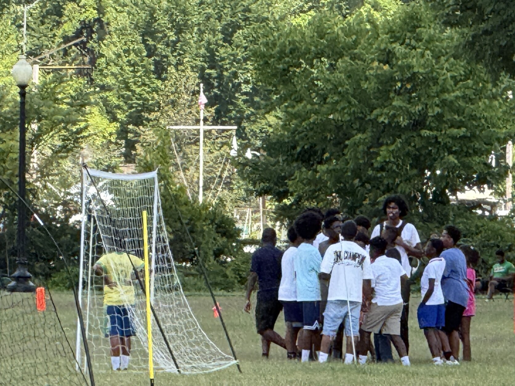 A crowd gathers to the left of a soccer net in a field, as a child in a green tee stands in the net