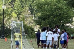 A crowd gathers to the left of a soccer net in a field, as a child in a green tee stands in the net