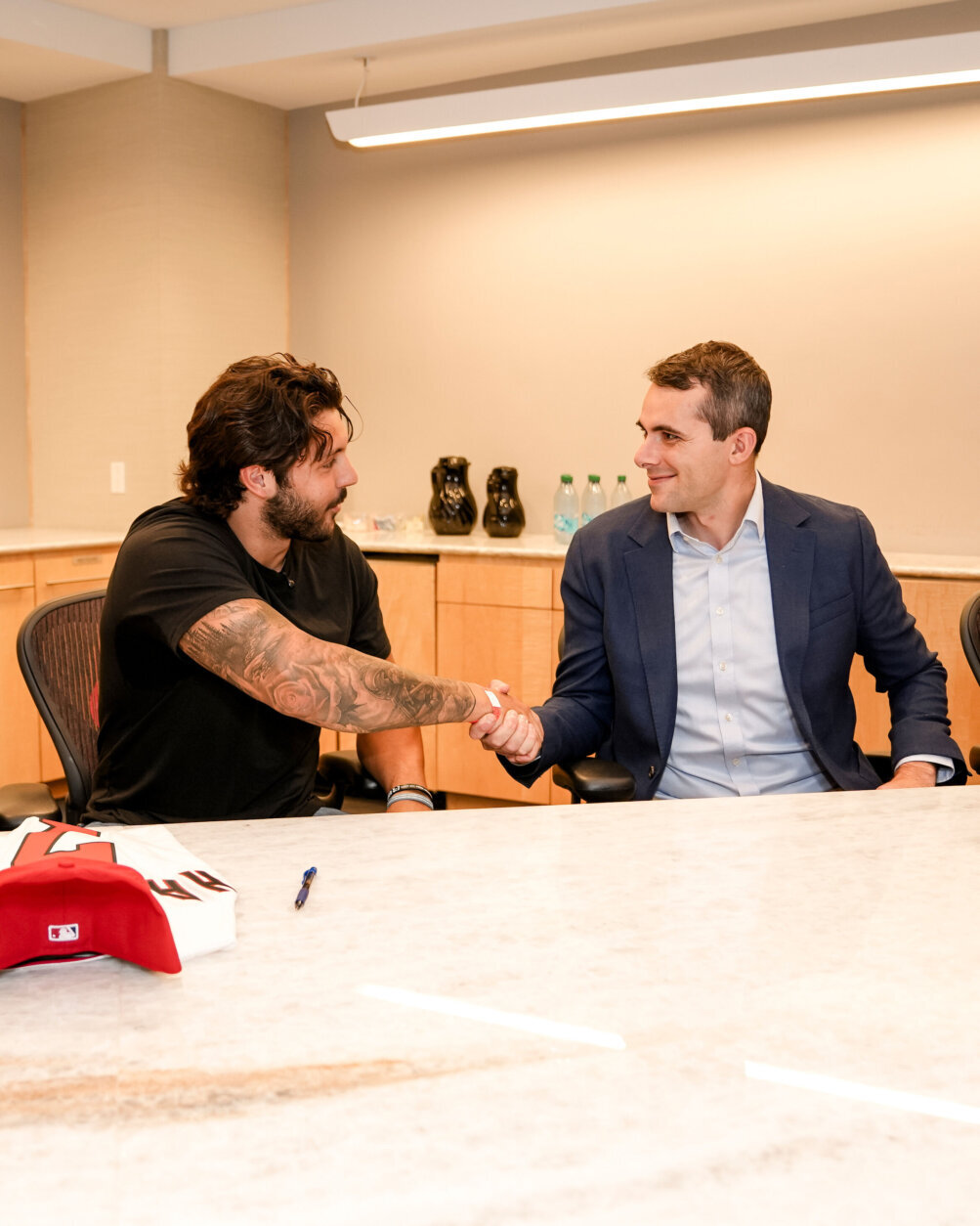 baseball player in street clothes on left shakes hand with business executive in room after signing contract