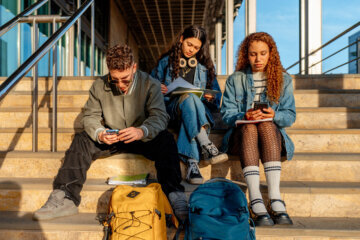 Three young adult university students are sitting on steps, using smartphones and studying, with backpacks beside them